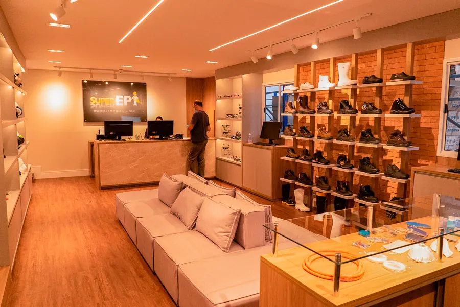 Interior de uma loja de calçados moderna com iluminação aconchegante, piso de madeira e prateleiras exibindo diversos modelos. Um cliente está em pé no balcão. Ambiente acolhedor e convidativo.